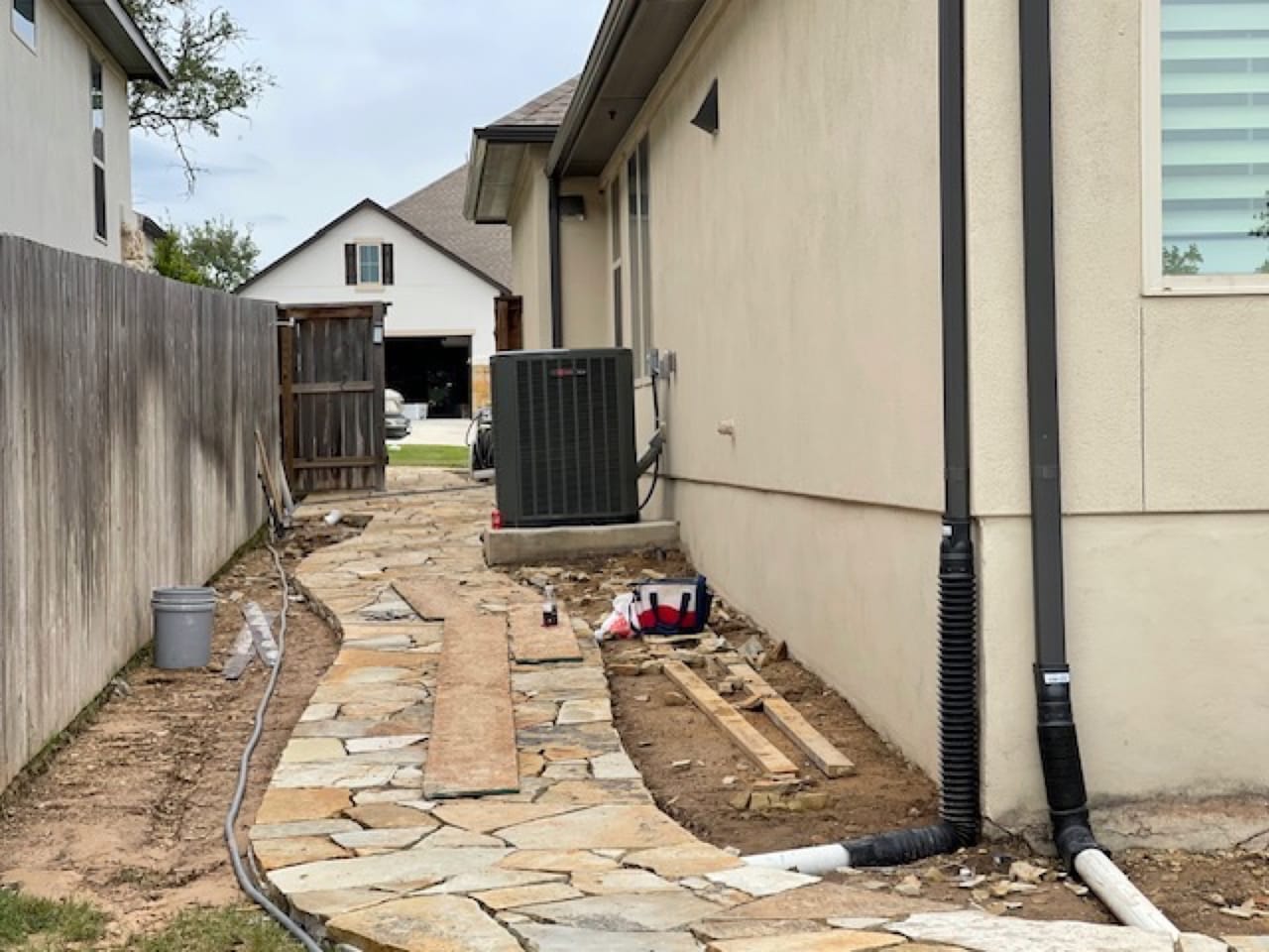 Narrow side yard with downspout drain pipes and partly laid flagstone path in Kyle, TX