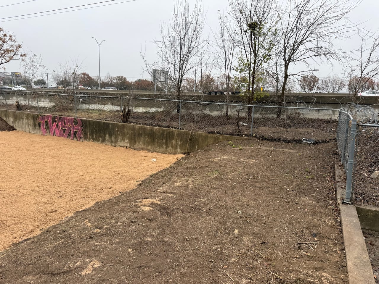 Bare graded dirt channel bottom between concrete walls with bare trees in Austin, TX