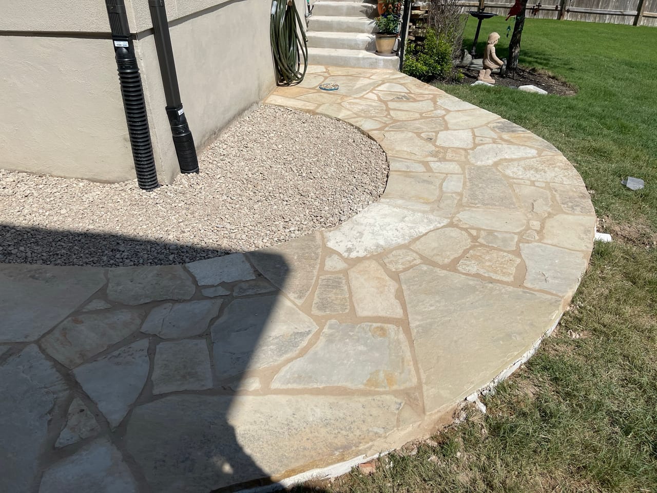 Curved flagstone patio edge meeting gravel, photographer shadow across frame by Corral Bros in Central Texas