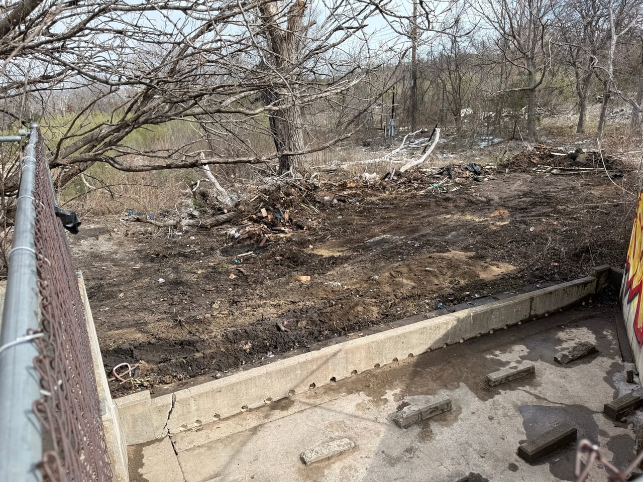 Cleared brush and dirt behind a concrete wall and chain link fence in Austin, TX