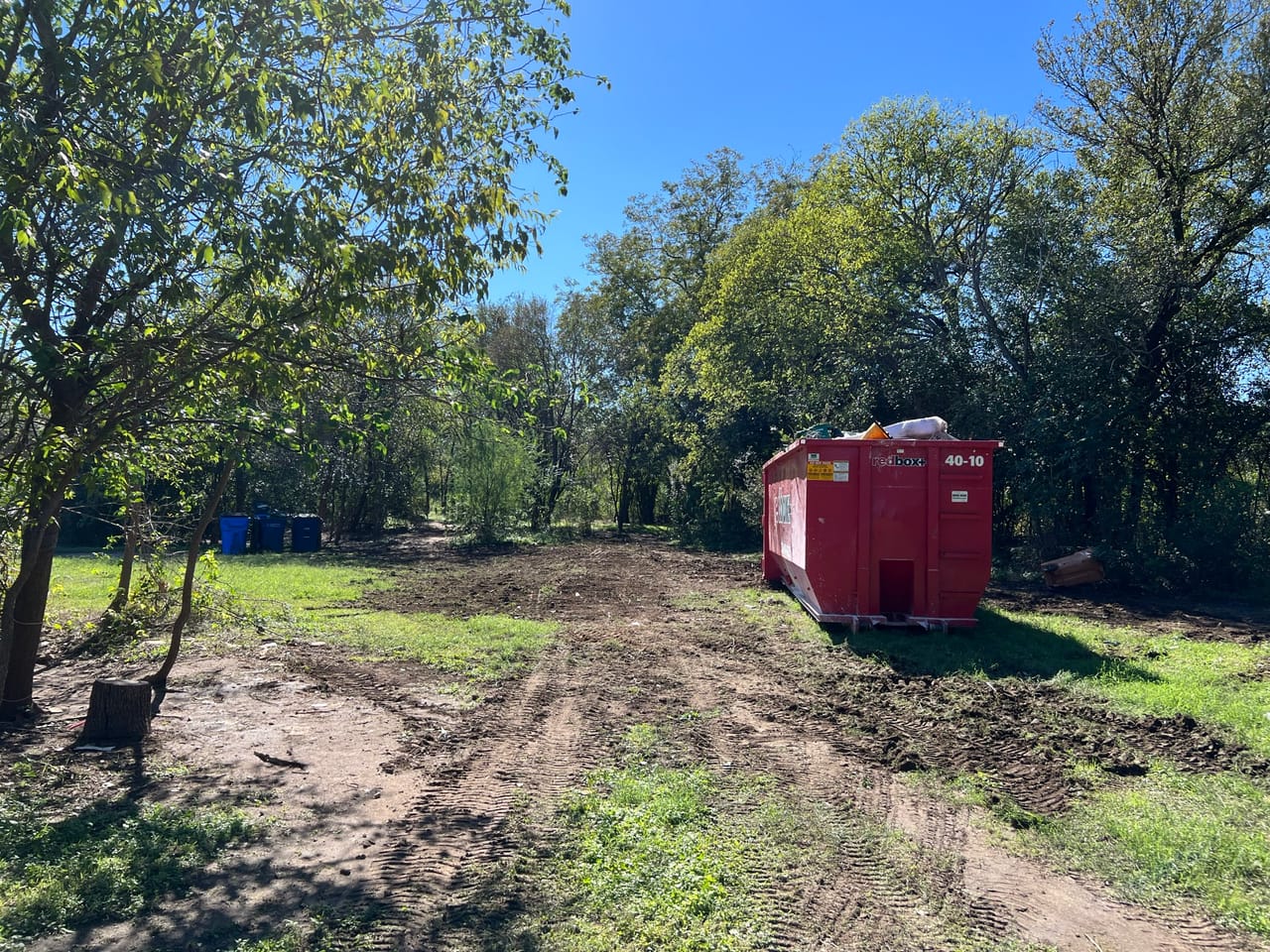 Cleared brushy lot with a red roll off dumpster and fresh tire ruts in Spicewood, TX
