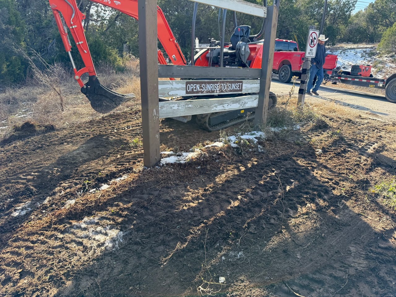 Excavator arm beside a wooden entrance sign on cleared tracked soil in San Marcos, TX