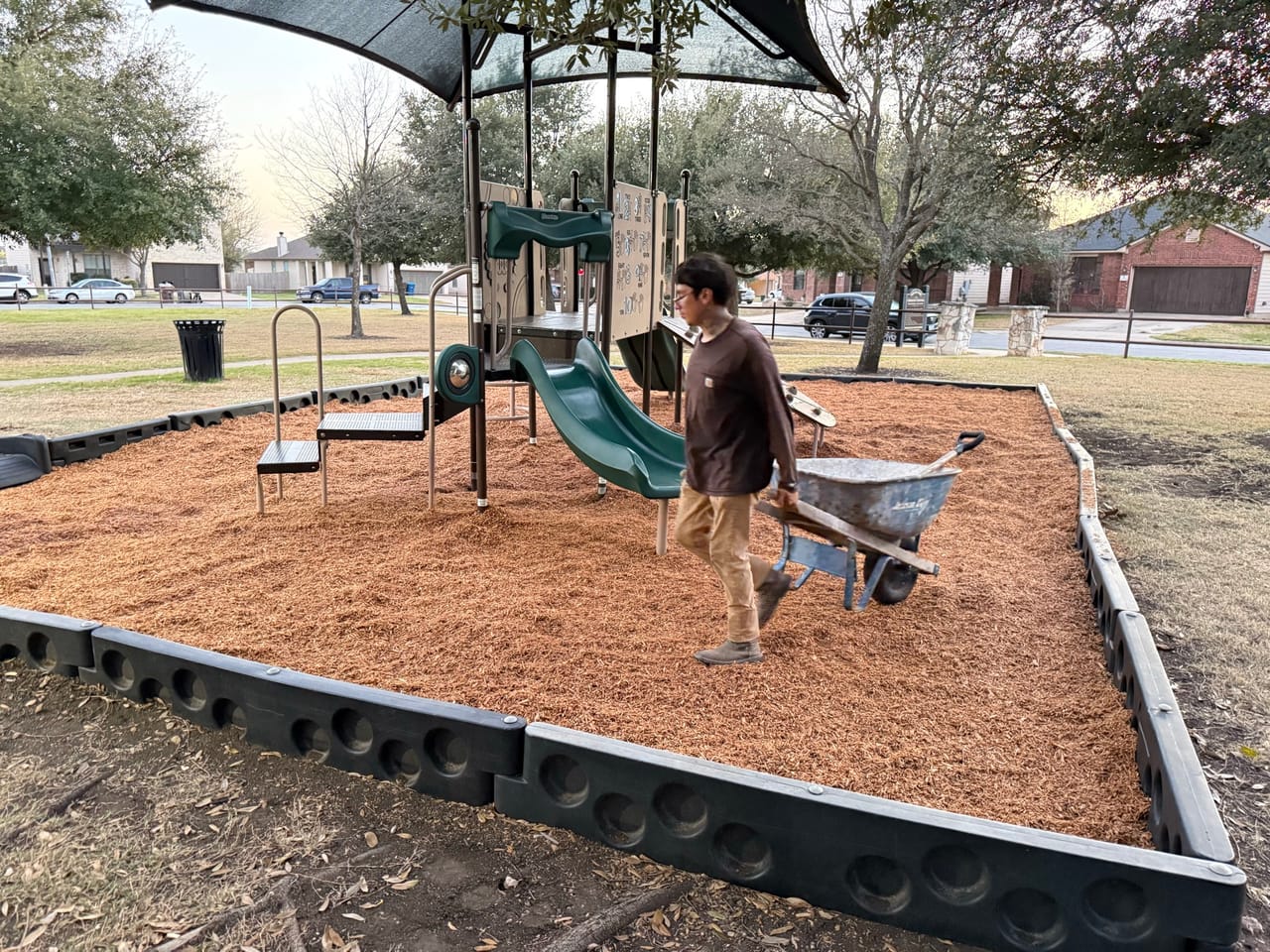 Worker hauling a wheelbarrow of mulch across a park playground in Kyle, TX
