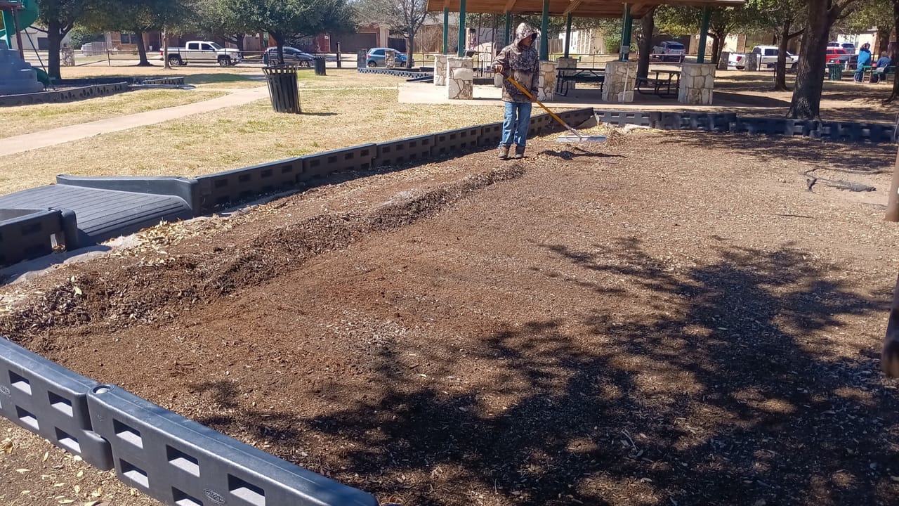 Worker raking fresh dark mulch into a wide bed at a park pavilion in Buda, TX