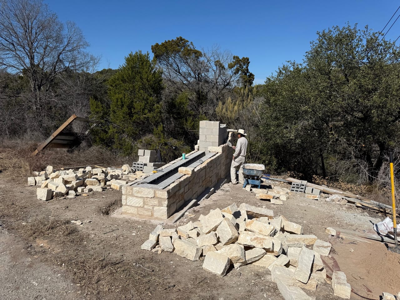 Mason working a limestone wall with cut stone piled around by Corral Bros in Central Texas