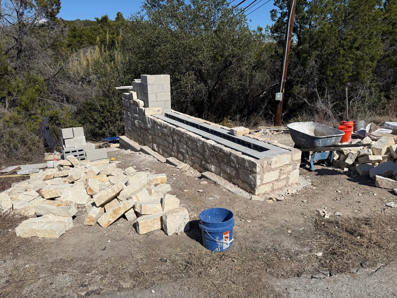 Stone veneer wall under construction with wheelbarrow and stacked blocks by Corral Bros in Central Texas
