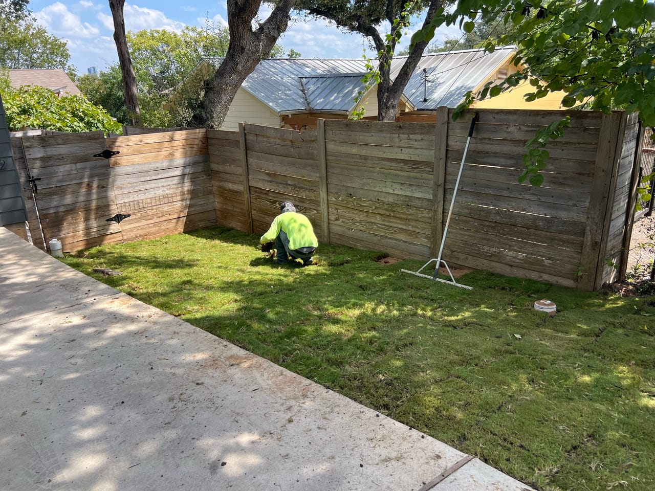 Worker in hi-vis kneeling on new sod lawn beside a fence by Corral Bros in Central Texas