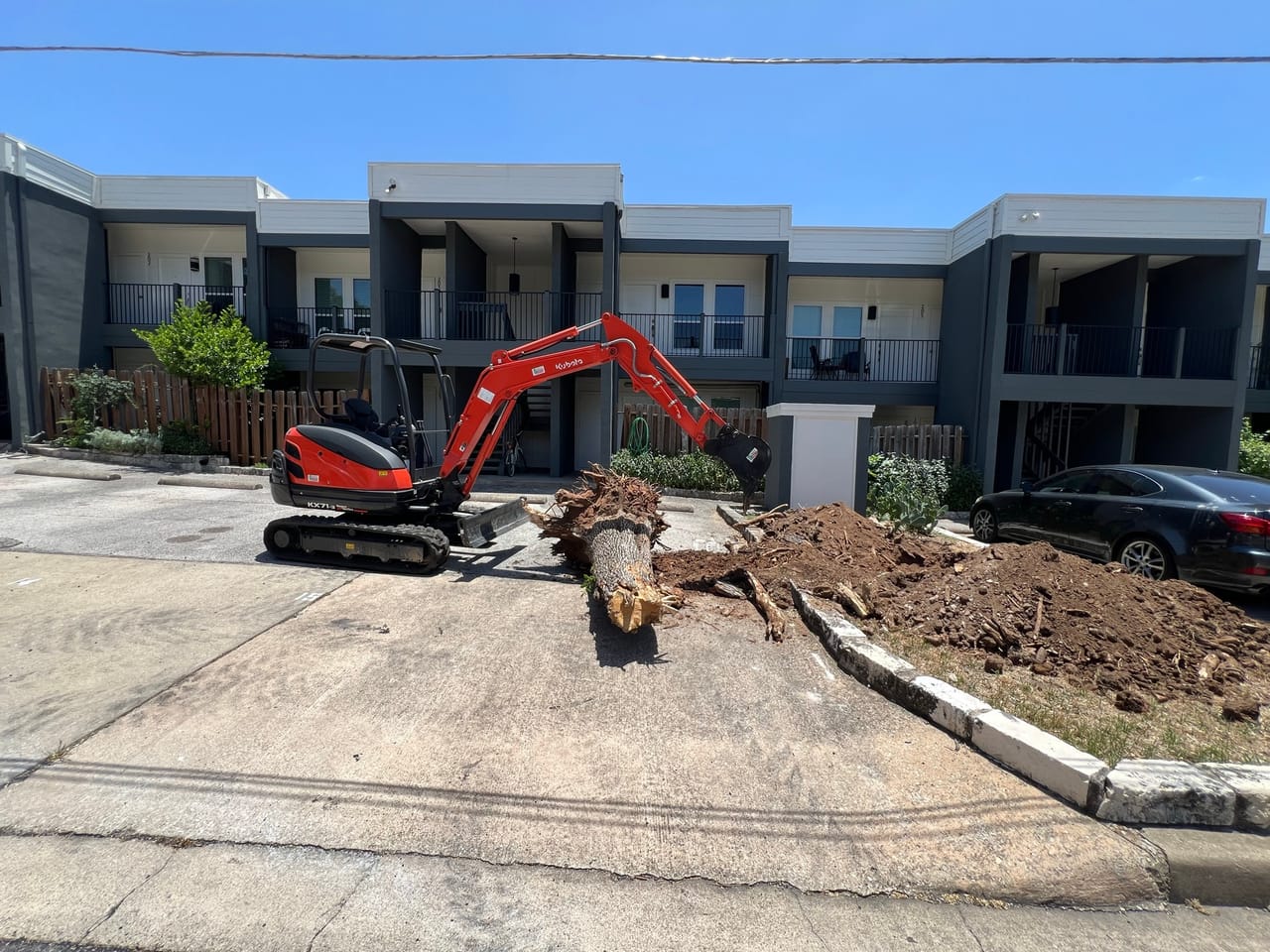 Red mini excavator pulling a stump out beside an apartment driveway in Leander, TX